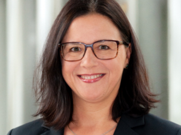 Cynthia Schönau Eine Frau mit langen, dunklen Haaren trägt eine Brille und ein schwarzes Blazer-Jacket. Sie lächelt freundlich in die Kamera, vor einem unscharfen hellen Hintergrund.