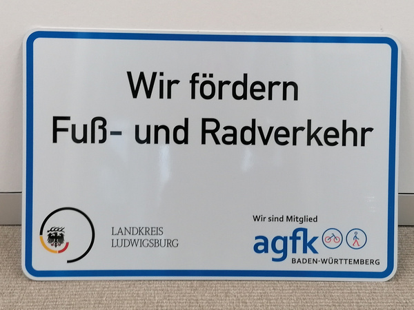 Ein Schild mit dem Text „Wir fördern Fuß- und Radverkehr“. Darüber steht das Logo des Landkreises Ludwigsburg und das Zeichen der AGFK Baden-Württemberg, einer Interessensgemeinschaft für Fuß- und Radverkehrsförderung.
