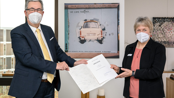 A man in a suit and a woman in business attire, both wearing masks, are exchanging documents in an office setting. A colorful artwork with the text "WE COME TOGETHER" is visible in the background. The atmosphere is formal, indicating a significant interaction, possibly an agreement or collaboration.
