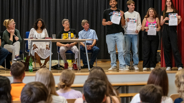 Eine Gruppe von Jugendlichen steht auf einer Bühne und hält Zertifikate in den Händen. Im Hintergrund sitzen mehrere Erwachsene an einem Tisch. Der Fokus liegt auf den strahlenden Gesichtern der Jugendlichen und ihrer Leistung.
