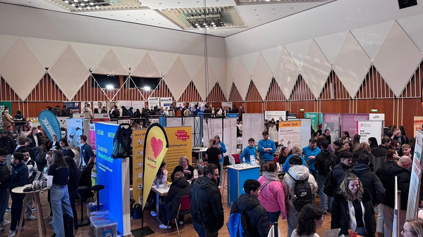 Eine Menschenmenge auf einer Messe in einem großen Raum, umgeben von bunten Informationsständen. Besucher interagieren mit Ausstellern und informieren sich über verschiedene Angebote. Die Atmosphäre ist geschäftig und lebhaft, mit vielen Teilnehmern, die Networking betreiben.