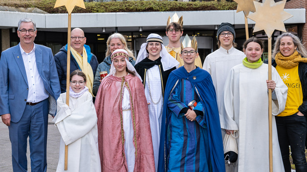 Eine Gruppe von Menschen steht in einem Innenhof. Sie tragen festliche Kleidung und halten Sterne in den Händen. Der Hintergrund zeigt ein modernes Gebäude. Die Atmosphäre ist fröhlich und feierlich, passend zu einer besonderen Veranstaltung oder Feier.