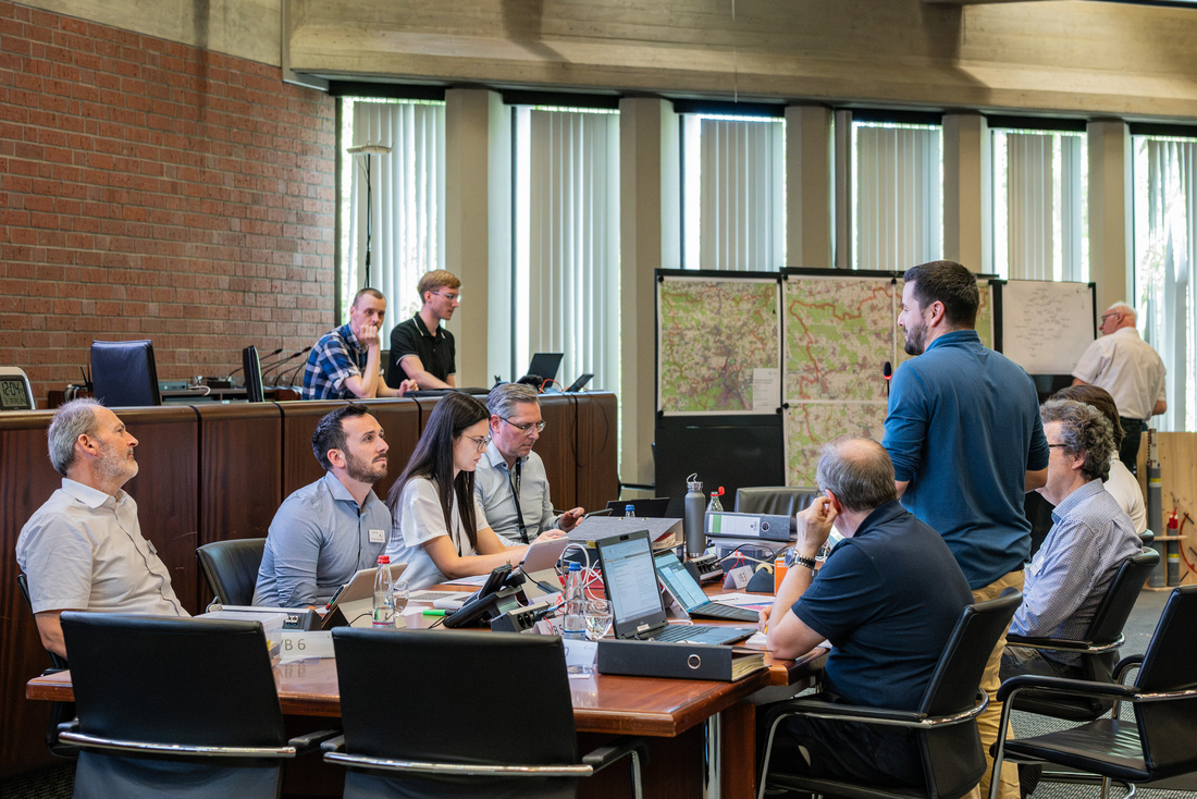Eine Gruppe von Fachleuten diskutiert an einem Konferenztisch, während sie Laptops und Dokumente nutzen. Einige Personen sitzen, andere stehen und sind in angeregtem Austausch. Im Hintergrund sind große Karten und Fenster mit Tageslicht zu sehen.