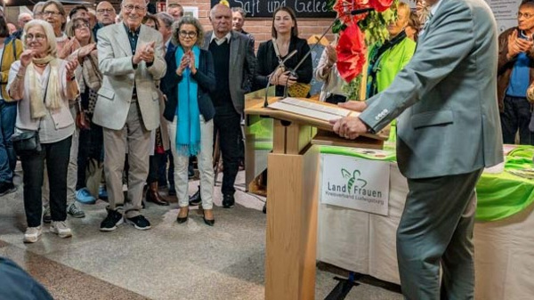 Ein Mann steht an einem Rednerpult und spricht zu einer Gruppe von Menschen, die applaudieren. Die Szene scheint eine Veranstaltung oder Feier zu sein, mit dekorativen Elementen im Hintergrund. Die Anwesenden wirken aufmerksam und interessiert.