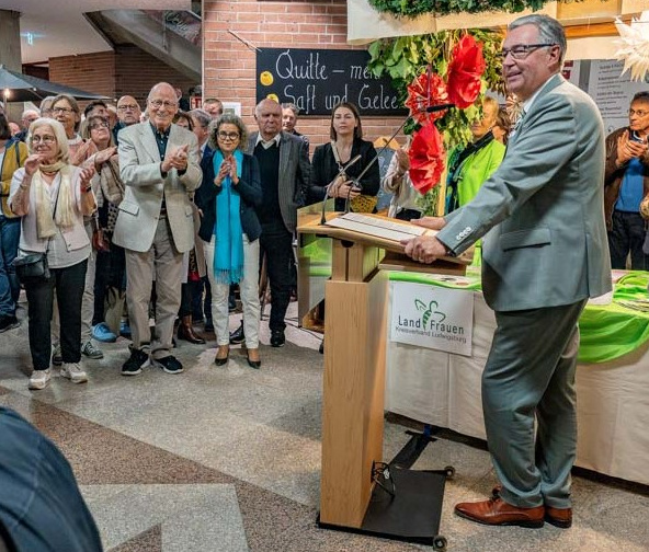 Ein Mann steht an einem Rednerpult und spricht zu einer Gruppe von Menschen, die applaudieren. Die Szene scheint eine Veranstaltung oder Feier zu sein, mit dekorativen Elementen im Hintergrund. Die Anwesenden wirken aufmerksam und interessiert.