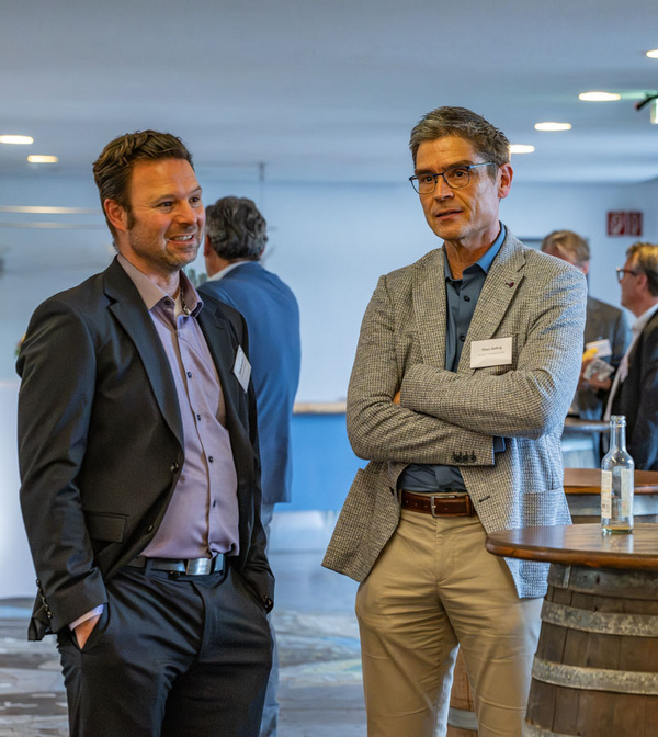 Zwei Männer stehen in einem modernen Veranstaltungsraum. Der links trägt einen schwarzen Anzug und hat eine entspannte Haltung, während der rechts in einem grauen Blazer mit verschränkten Armen steht. Im Hintergrund sind weitere Personen zu sehen, die miteinander sprechen.