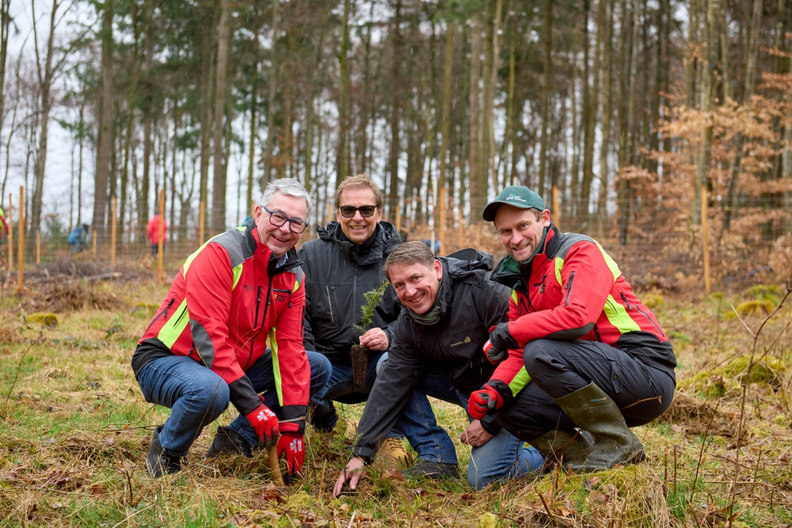 Vier Männer in Arbeitskleidung knien auf einem Waldboden und pflanzen gemeinsam einen Baum. Sie lächeln in die Kamera und wirken engagiert. Im Hintergrund sind verschwommen weitere Personen und Bäume sichtbar.