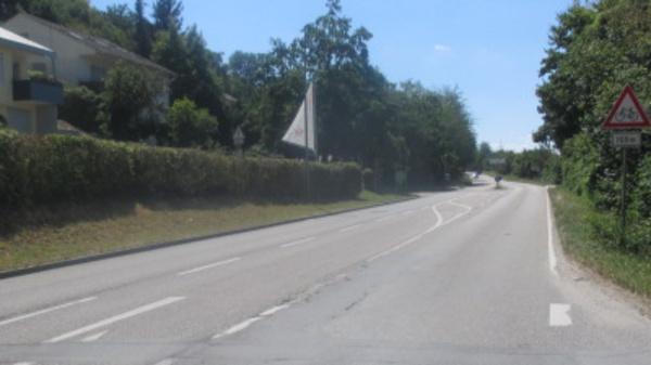 A view of a curved road surrounded by greenery. On the right, there are trees and bushes, while buildings are visible on the left. A road sign indicating caution is present, but the specific warning is not discernible. The sky is clear with a few clouds.