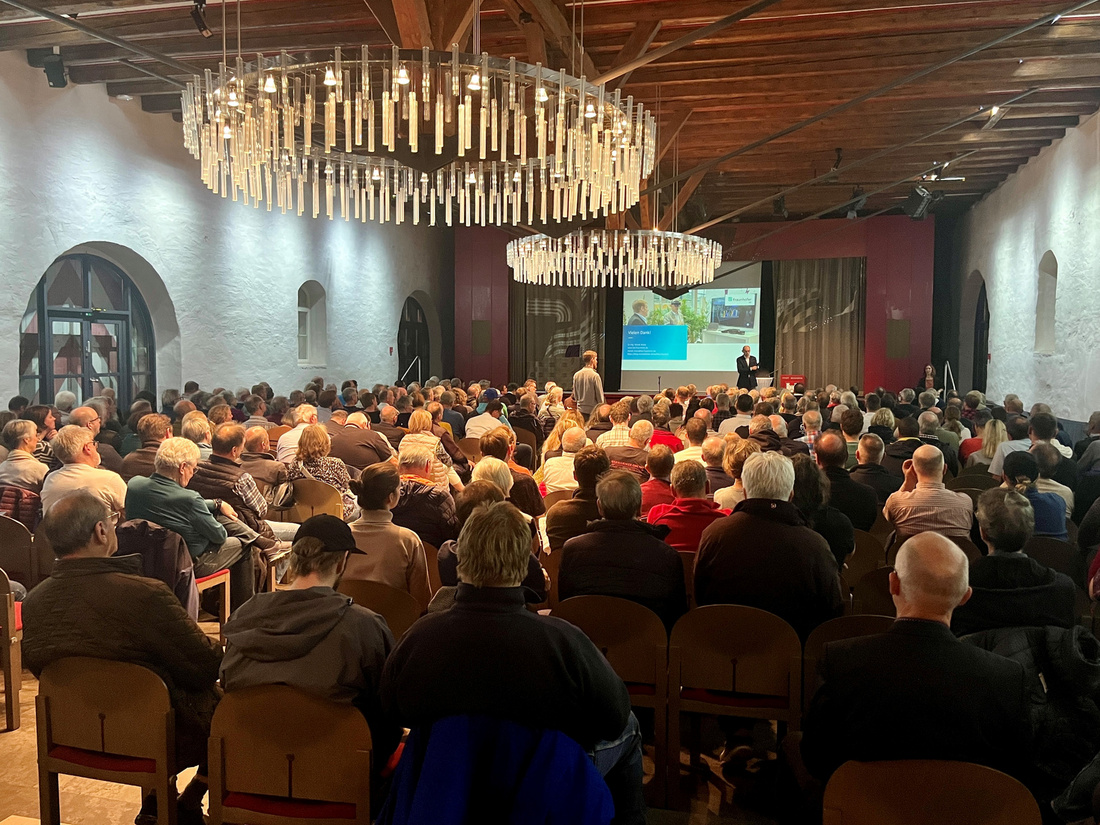 Ein voll besetzter Raum mit einer großen Anzahl von Menschen, die auf eine Präsentation auf einer Leinwand blicken. Eine Person steht auf der Bühne und spricht zum Publikum. Die Atmosphäre wirkt aufmerksam und interessiert.