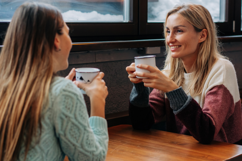 Two women are seated at a wooden table, enjoying warm beverages. They are engaged in a friendly conversation, smiling at each other. The setting features large windows with a view of snow, creating a cozy atmosphere.