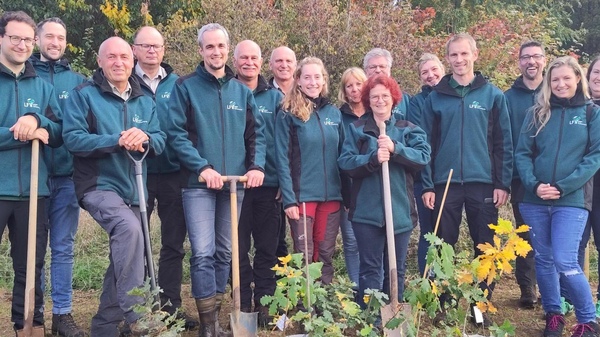 Un groupe de quinze personnes se tenant debout, vêtues de vestes vertes, sur un terrain en pleine nature. Elles tiennent des pelles et sont entourées de pots de plantes prêtes à être plantées. L'arrière-plan comprend des arbres aux couleurs d'automne.