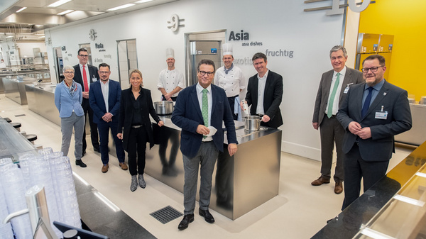 Eine Gruppe von zwölf Personen steht in einer modernen Küche, umgeben von Kochbereichen. Einige Personen tragen Kochjacken. Die Atmosphäre wirkt professionell und freundlich, als ob ein Event oder eine Eröffnung stattfindet. Im Hintergrund sind Beschriftungen zu sehen, die verschiedene Küchenstile angeben.