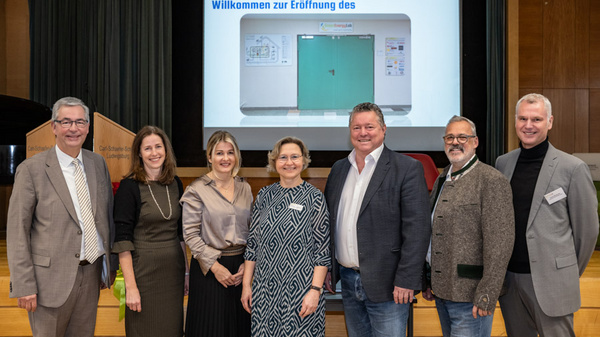 Eine Gruppe von sieben Personen steht auf der Bühne vor einem Bildschirm, auf dem "Willkommen zur Eröffnung des" mit einem Bild einer grünen Tür zu sehen ist. Die Personen lächeln und tragen formelle Kleidung.