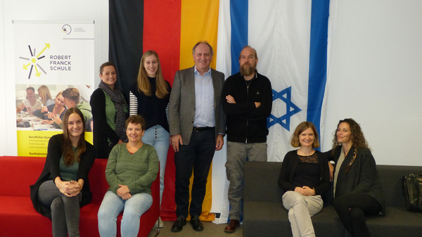 Grupp portrait in einem Schulungsraum, in dem sieben Personen stehen und sitzen. Im Hintergrund sind die Flaggen Deutschlands und Israels zu sehen, sowie ein Schild der Robert Franck Schule. Alle Personen lächeln und scheinen an einem gemeinsamen Projekt beteiligt zu sein.