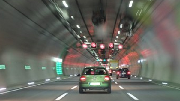 Ein Tunnel mit mehreren Fahrspuren. Ein grünes Auto fährt in Richtung der roten Ampeln. Die Wände des Tunnels sind beleuchtet, und es gibt grüne Hinweisschilder. Die Tunnelbeleuchtung schafft eine dynamische Atmosphäre, während die Fahrzeuge sich auf der Straße bewegen.