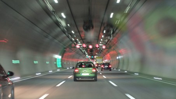 Ein Tunnel mit mehreren Fahrspuren. Ein grünes Auto fährt in Richtung der roten Ampeln. Die Wände des Tunnels sind beleuchtet, und es gibt grüne Hinweisschilder. Die Tunnelbeleuchtung schafft eine dynamische Atmosphäre, während die Fahrzeuge sich auf der Straße bewegen.