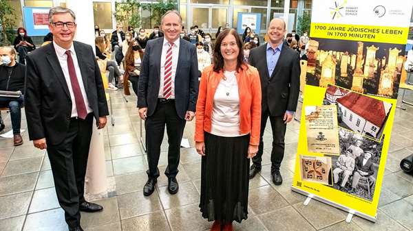 Quatre personnes se tiennent debout dans un espace public, souriant et vêtues de costumes formels. À droite, une affiche colorée présente un projet sur la vie juive, intitulé « 1700 Jahre jüdisches Leben in 1700 Minuten ». En arrière-plan, un groupe de personnes assises regarde.