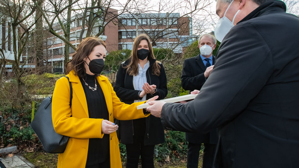 Eine Person mit gelber Jacke erhält von einem Mann ein Dokument. Im Hintergrund stehen zwei Frauen und ein Mann. Alle Personen tragen Masken. Die Szene spielt sich im Freien vor einem Gebäude ab.