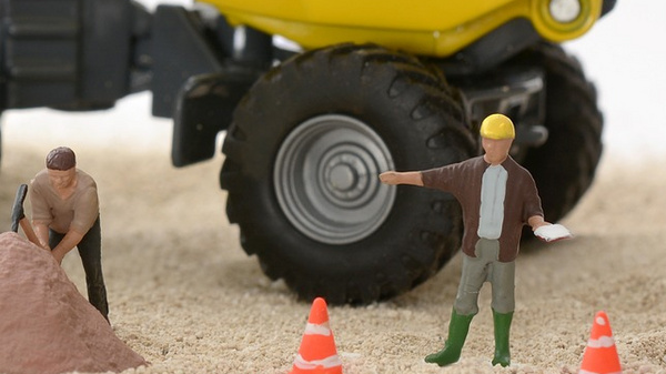 A miniature construction scene features a large yellow dump truck and three figurines: one uses a wheelbarrow, another stands beside a pile of dirt, and a third holds a clipboard. Surrounding the scene are miniature traffic cones and construction signs. Sand serves as the ground.