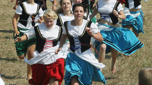 A group of women in traditional dresses run energetically through a field. Their outfits include vibrant skirts and white blouses, showcasing a festive atmosphere. The scene captures a sense of excitement and community as they participate in a spirited event.