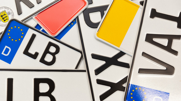Mehrere Autokennzeichen liegen übereinander, darunter sind die Buchstabenkombinationen "LB" und "VAI" sowie europäische Abzeichen zu sehen. Einige der Schilder haben unterschiedliche farbige Rahmungen in Rot und Gelb. (Text KI generiert)