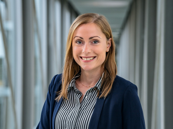 Eine lächelnde Frau mit langen, blonden Haaren steht in einem modernen Innenraum mit großen Fenstern. Sie trägt ein dunkles Blazerjackett und ein gestreiftes Hemd. Ihr freundlicher Ausdruck vermittelt Offenheit und Professionalität.