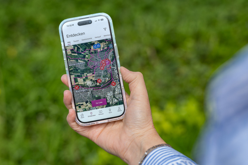 Eine Hand hält ein Smartphone, auf dessen Bildschirm eine Kartenansicht mit verschiedenen markierten Punkten zu sehen ist. Der Hintergrund zeigt grünes Gras. Die App scheint Funktionen zum Erkunden einer Umgebung anzubieten.