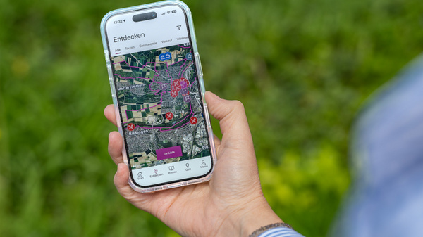 Eine Hand hält ein Smartphone, auf dessen Bildschirm eine Kartenansicht mit verschiedenen markierten Punkten zu sehen ist. Der Hintergrund zeigt grünes Gras. Die App scheint Funktionen zum Erkunden einer Umgebung anzubieten.