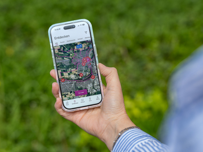 Eine Hand hält ein Smartphone, auf dessen Bildschirm eine Kartenansicht mit verschiedenen markierten Punkten zu sehen ist. Der Hintergrund zeigt grünes Gras. Die App scheint Funktionen zum Erkunden einer Umgebung anzubieten.