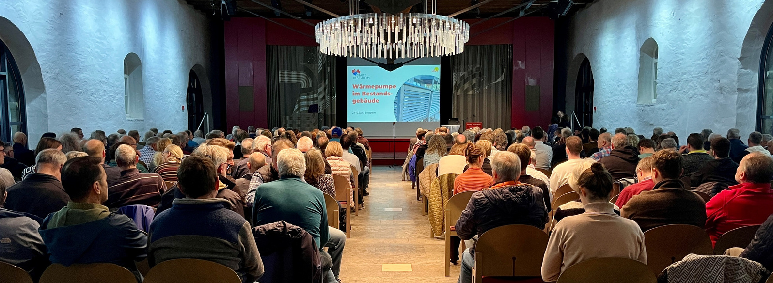 Header Bild: Ein voll besetzter Veranstaltungssaal mit vielen Personen, die auf eine Präsentation auf der Leinwand schauen. Die Teilnehmer sitzen in Stuhlreihen, während im Vordergrund ein Sprecher spricht. Die Beleuchtung ist angenehm und der Raum hat eine moderne Ausstattung.