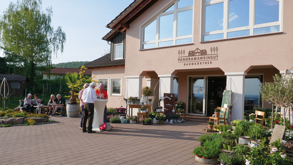 Auf dem Bild ist ein Weingut mit einer Terrasse zu sehen. Vor dem Gebäude stehen Tische und Pflanzen, während Gäste im Freien sitzen und sich unterhalten. Die Fassade des Weinguts ist freundlich und einladend gestaltet, mit großen Fenstern und dem Schriftzug „Panoramawinzer Baumgärtner“.