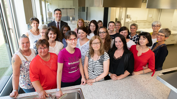 A group of around 20 women, along with a man, stand together in a modern kitchen setting. They are smiling and appear to be engaged in a community or team activity. The atmosphere is friendly and collaborative.