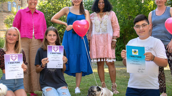 Eine Gruppe von sieben Personen steht in einem Garten. Einige halten Luftballons und Zertifikate. Vorne sitzen zwei Hunde. Die Umgebung wirkt freundlich und festlich, mit einem Gebäude im Hintergrund, das zur Szene beiträgt.