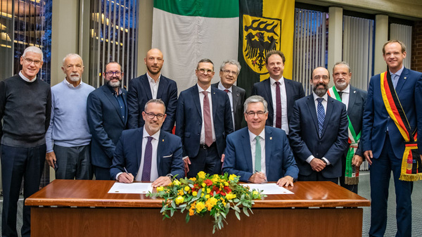 07.11.2022 - Unterzeichnung der Partnerschaftsurkunde mit der Provinz Bergamo Eine Gruppe von Männern steht um einen Tisch, an dem zwei Männer sitzen und Dokumente unterschreiben. Im Hintergrund hängen Flaggen. Die Szene vermittelt den Eindruck eines offiziellen Ereignisses, möglicherweise einer Vereinbarung oder Unterzeichnung.