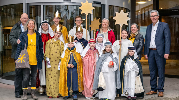 Un groupe de personnes, dont des enfants déguisés en rois mages, pose devant un bâtiment. Ils portent des costumes colorés et tiennent des étoiles en carton. L'ambiance est festive, célébrant une tradition culturelle.