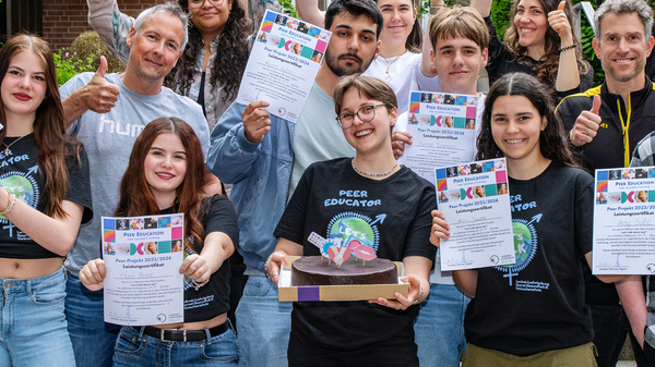 Eine Gruppe von Personen steht vor einer Gebäude, lächelt und zeigt Daumen nach oben. Sie halten Zertifikate in den Händen und feiern offensichtlich einen Erfolg oder Abschluss. Die meisten tragen schwarze T-Shirts mit verschiedenen Motiven. Die Stimmung ist positiv und enthusiastisch.