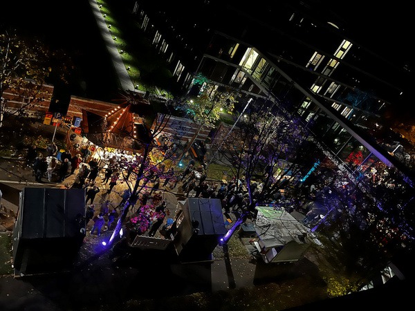 Eine Luftaufnahme einer belebten Veranstaltung bei Nacht. Menschenmengen bewegen sich zwischen bunten Ständen und Lichtern, während umgebende Bäume und Gebäuden angestrahlt werden. Die Atmosphäre ist festlich und lebendig.