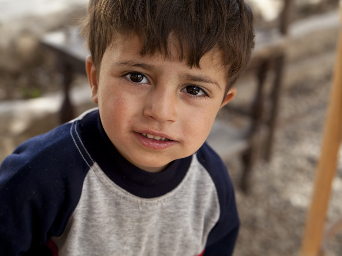 Bild: MaximilianV, Lizenz: CC BY 2.0 Ein junger Junge mit braunen Haaren schaut in die Kamera. Er trägt ein graues und dunkelblaues Sweatshirt. Der Hintergrund zeigt unscharfe Holzstühle und einen natürlichen Boden. Sein Gesichtsausdruck wirkt neugierig und freundlich.