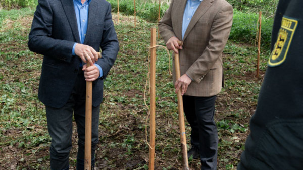 Two men stand in a forested area, each holding a shovel. They are dressed casually and appear engaged in a planting or gardening initiative. The background features green trees and ground cover, indicating an effort to enhance the natural environment.