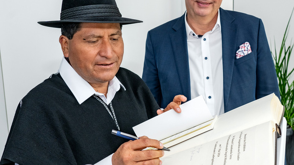 Ein Mann in traditioneller Kleidung unterschreibt ein Dokument, während ein anderer Mann in formeller Kleidung daneben steht und zuschaut. Der Raum hat eine moderne Atmosphäre mit einigen Pflanzen im Hintergrund.