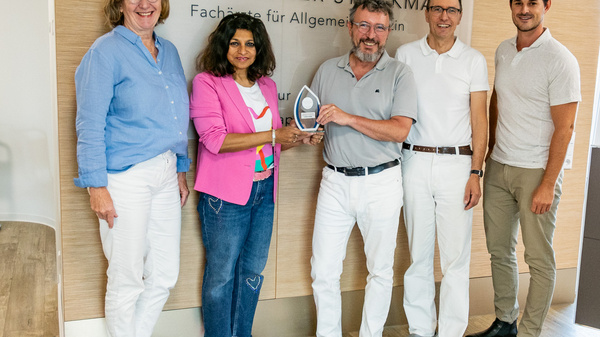 Gruppenbild von fünf Personen in einer Praxis. Sie stehen zusammen und lächeln. Eine Person hält einen Preis oder eine Auszeichnung in der Hand. Im Hintergrund ist ein Schild mit den Namen Dr. Axel Stöckmann und Dr. Rainer Stöckmann zu sehen, sowie die Beschreibung „Facharzt für Allgemeinmedizin“.