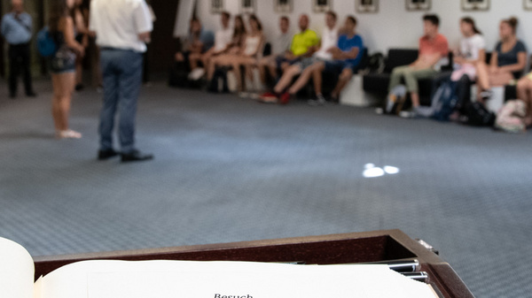 An open guestbook is in the foreground, displaying a message about a visit by a Hungarian delegation related to a youth protection camp on July 31, 2019. In the background, a group of people is seated, with someone speaking to them.