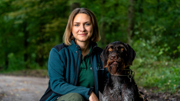 A woman with shoulder-length brown hair, wearing a dark jacket and green shirt, sits beside a brown and black dog in a forested area. They both look towards the camera, conveying a friendly and calm bond. The surroundings are lush with green trees.