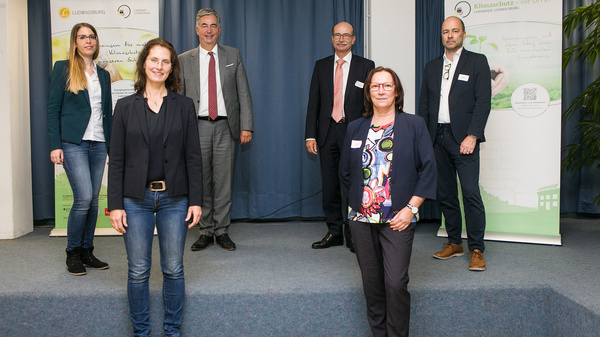 Cinq personnes se tiennent sur une scène, entourées de bannières. Une femme au premier plan porte un blazer noir et est flanquée de quatre hommes en arrière-plan, arborant des vêtements formels. Tous semblent participer à un événement professionnel.