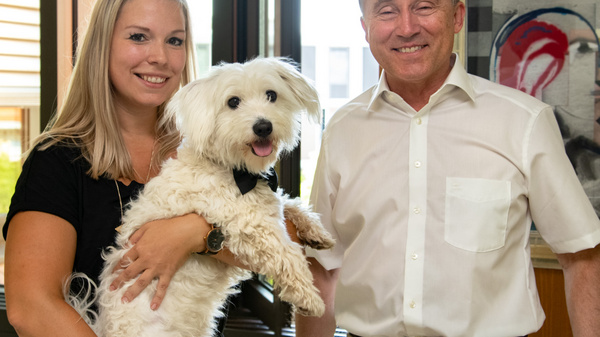 A woman holds a small dog while standing next to a man. They are both smiling, with a modern office environment in the background. The scene conveys a sense of warmth and companionship between the people and the dog.