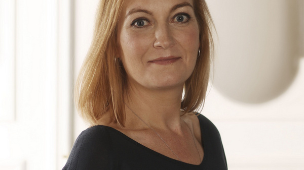 Ritratto di una donna con capelli castani lunghi e lisci, che indossa un maglione scuro. È in piedi e guarda verso la macchina fotografica, con un'espressione serena e sicura. Lo sfondo è chiaro, creando un contrasto con il suo abbigliamento.