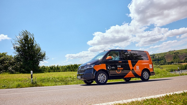 Un véhicule aux couleurs orange et noir, marqué "Rider", roule le long d'une route bordée d'herbe et d'arbres. Le ciel est bleu avec quelques nuages. Le véhicule semble être en mouvement, illustrant un service de transport.
