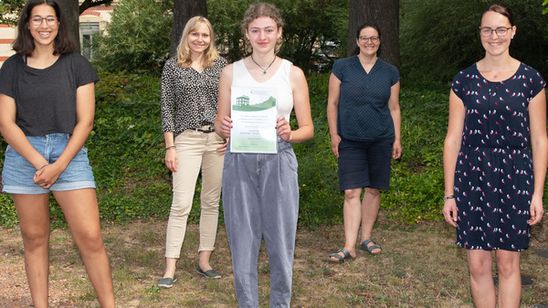 A group of seven people stands outdoors in a grassy area, surrounded by trees. They are posing together, with one person holding a document in the front. The group includes a mix of genders and attire, with some in casual clothing and others in business attire.