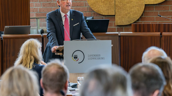 Ein Mann in Anzug spricht am Pult vor einem Publikum in einem Sitzungssaal. Im Hintergrund ist ein großes Kunstwerk eines Adlers sichtbar. Die Zuhörer sitzen hinter ihm und folgen aufmerksam seiner Rede.