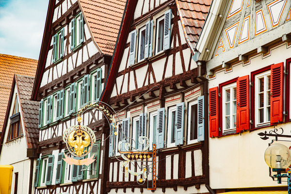 Die Abbildung zeigt ein traditionelles Fachwerkhaus mit einem roten und grünen Fensterladen. An der Fassade ist ein dekoratives Schild angebracht. Die Architektur ist typisch für viele historische Städte in Deutschland und vermittelt ein rustikales, charmantes Ambiente.